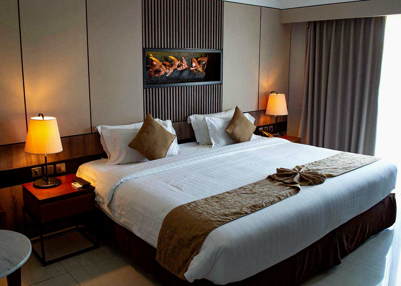 A warm room with two reading lamps, a long bed and a dark panelled headboard